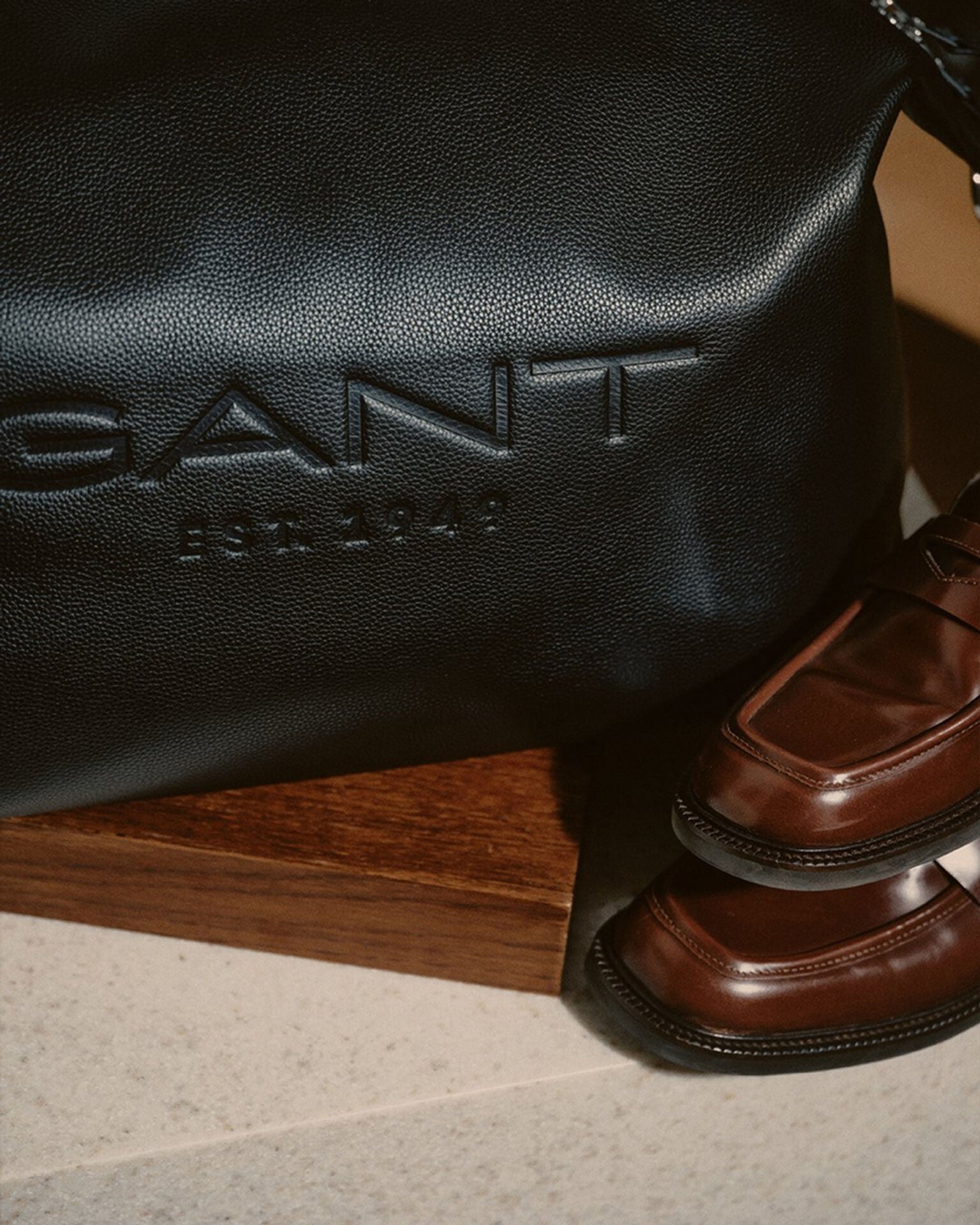 Black leather bag with 'Gant' branding next to a brown leather shoe on a wooden surface.