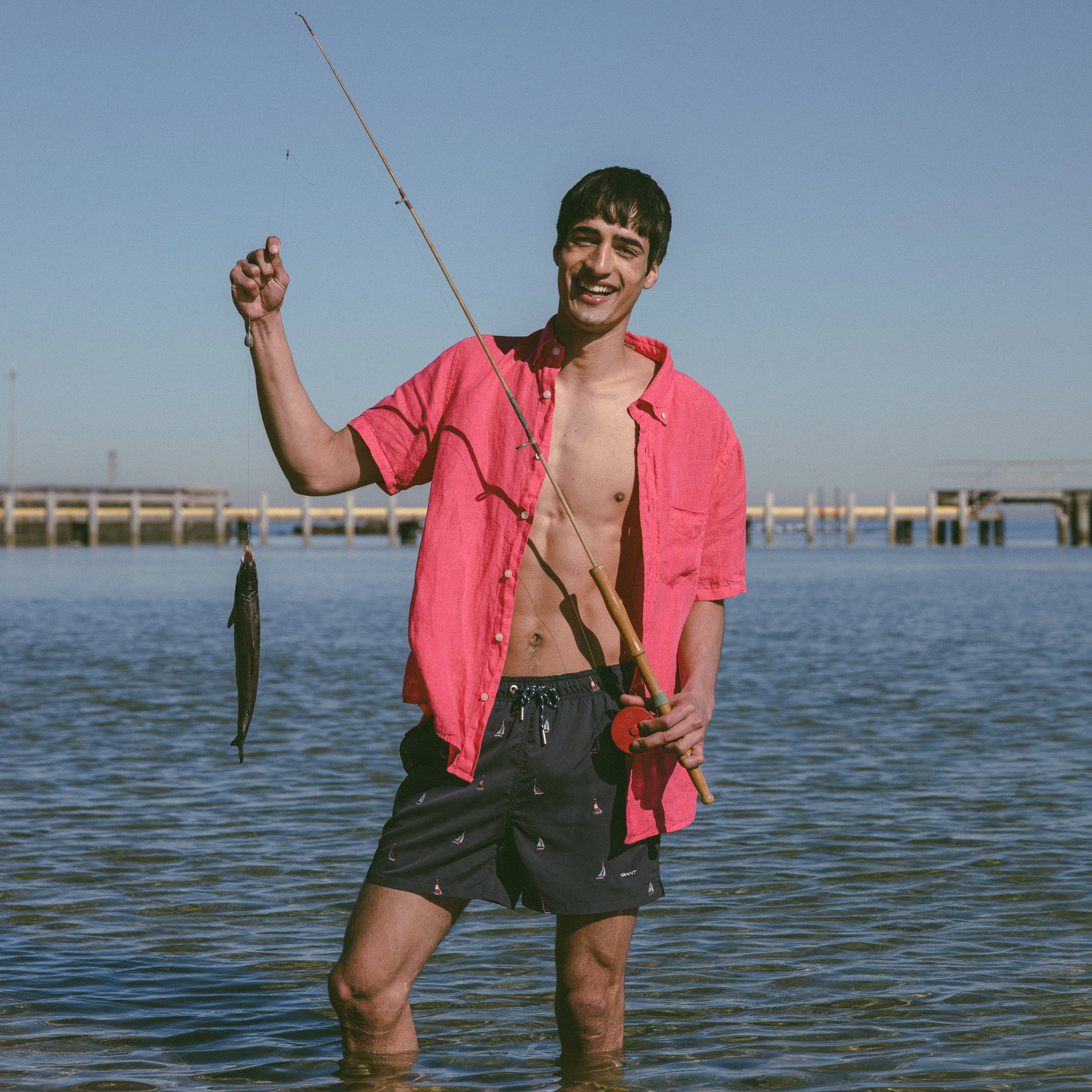 Man in GANT short sleeve linen shorts and swim shorts holding a fishing rod and fish in water with pier in background