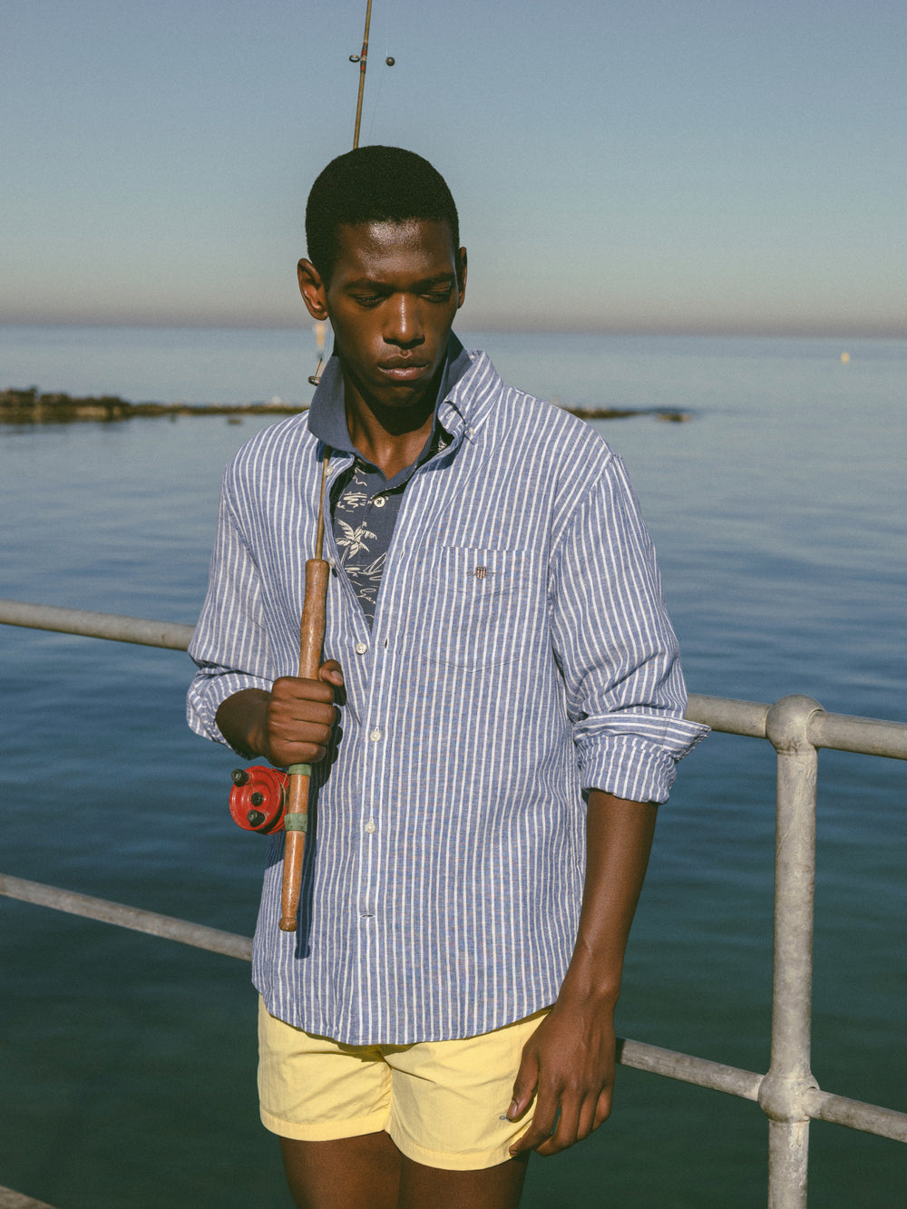 Man holding a fishing rod by the water, wearing the GANT Regular Fit Striped Cotton Linen Shirt in rich blue.